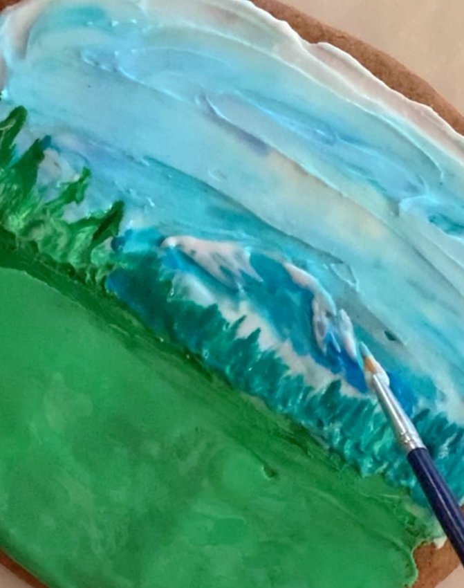 Spread and blend icing on the bottom half of the cookie to make the field. Add mountains to the top half using a palette knife and a brush. Add color to the foreground with a small brush, using a horizontal motion.